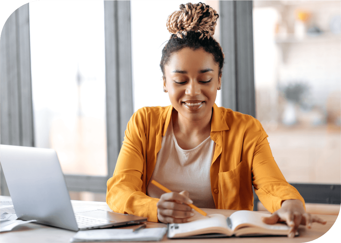 Young woman sitting at desk with notebook, pencil and laptop smiling at her work.