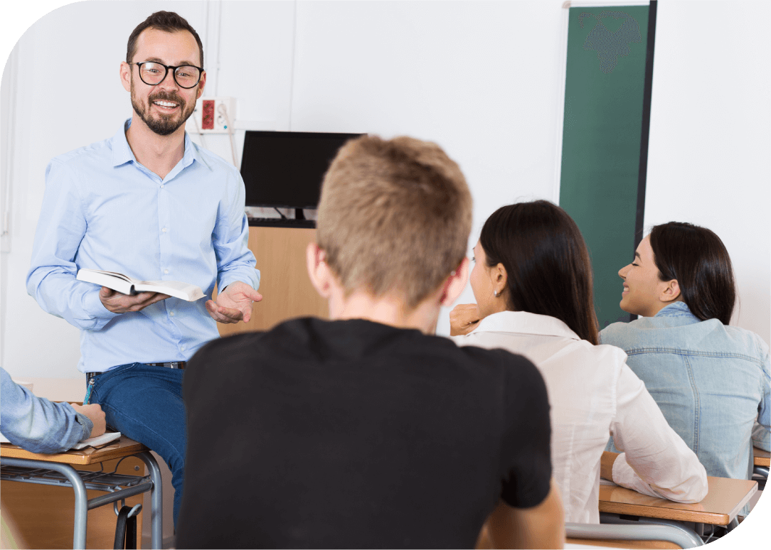 Male class instructor standing in classroom holding book and leaning against desk talking to adult students.