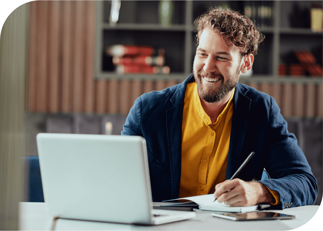 Man sitting at desk with notebook, pencil and laptop smiling at his screen.